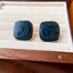 Detail of dark green square enamel earrings with gold edges on a beige velvet cushion