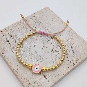 Close-up of pink evil eye charm on golden beaded bracelet with adjustable woven cord
