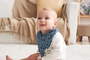 Baby boy wearing a blue leopard print muslin bib sitting on a beige carpet at home
