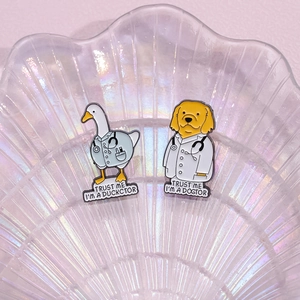 Cartoon enamel pins of duck and dog doctors on an iridescent shell tray