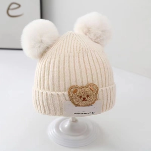 Cozy cream baby knit hat featuring dual pom-poms and a cute cartoon bear patch