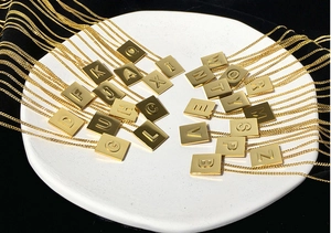 Golden titanium steel square alphabet pendants displayed on a white tray with black background