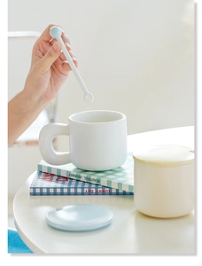 Close-up of a white ceramic mug set with large round handle, lid and spoon in use