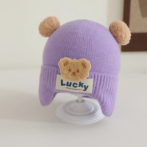 Close-up of light purple knitted hat with plush bear patch and double pom-poms details