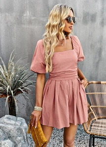 Pink square-neck romper with waist tie and pleated hem shown in a casual lifestyle setting