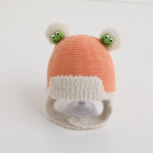 Close-up of baby knit earflap hat with orange yarn, white plush trim and frog pom-poms