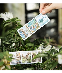 Close-up of watercolor scenery stickers held by hand among green leaves and white flowers