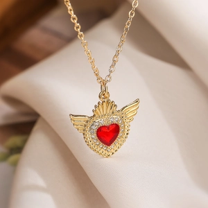 Gold plated heart pendant necklace with red gemstone and wings on beige fabric