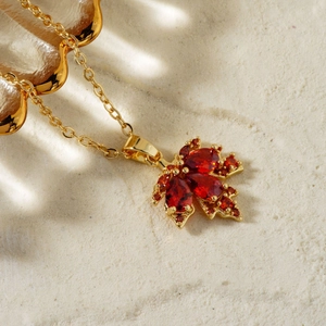 Collana a foglia d'acero dorata con zirconi rossi su sfondo bianco morbido