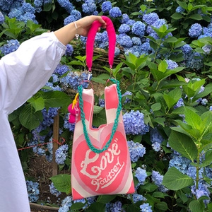 Pink knit tote bag with heart pattern and green chain held against blue hydrangeas