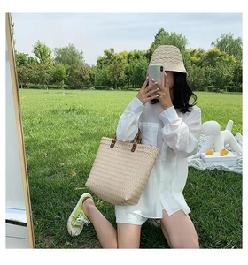 Detailed mirror selfie of a beige woven straw tote bag with dark handles on a grass field