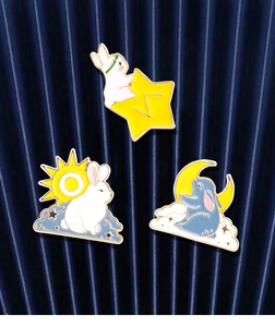 Three enamel bunny brooches with sun, moon and star motifs on dark blue fabric background