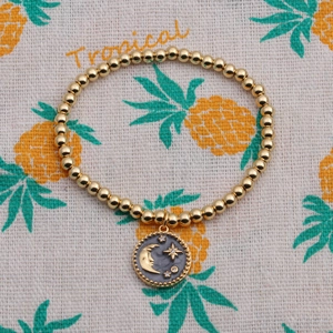 Detail von goldenem Perlenarmband mit Mond- und Sternanhänger auf tropischem Hintergrund