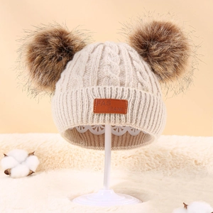 Detailed close-up of knitted beige baby hat with dual brown faux fur pom poms