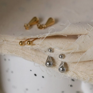 Orecchini minimalisti a goccia in oro e argento presentati su tessuto di lino testurizzato