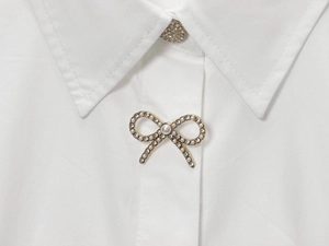 Dettaglio di spilla a fiocco dorata con perle e strass sul colletto di camicia bianca