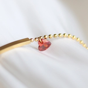 Macro detail of 18K gold-plated beaded bracelet with red heart crystal and bar charm