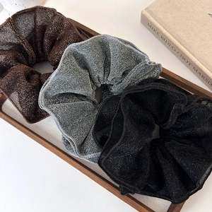 Three shimmering double-layered scrunchies in brown, silver, and black on a wooden tray