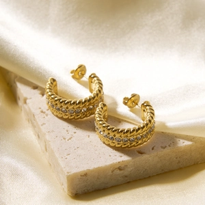 Detail of gold C-shaped twisted hoop earrings with sparkling zircon inlay on stone surface