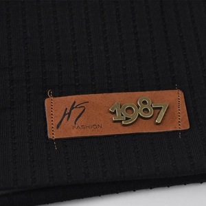 Primo piano di un berretto in maglia nera con toppa in pelle marrone e logo metallico 1987.