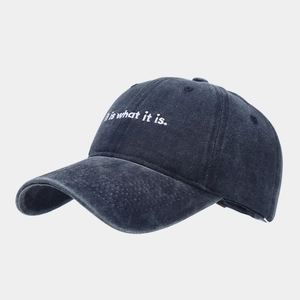 Dettaglio del ricamo bianco 'it is what it is' sul cappello da baseball in denim