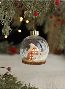 Detalle de bola de Navidad LED con cabaña de madera y reno en rama de pino