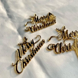 Three pieces of golden acrylic Merry Christmas cake toppers on a white textured background