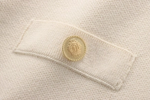 Close-up of cream knit fabric featuring a decorative gold crest button and pocket flap detail