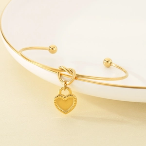 Minimalist gold stainless steel knotted bangle with heart-shaped pendant on white tray