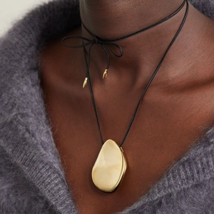 Detail of an irregular golden pendant with a black cord and metallic end tips on a model