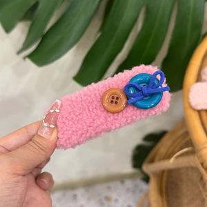 Handheld pink plush hair clip with buttons and a blue bow detail, soft aesthetic