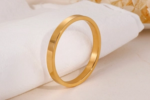 Vertical view of a solid gold-toned polished stainless steel bangle on white fabric