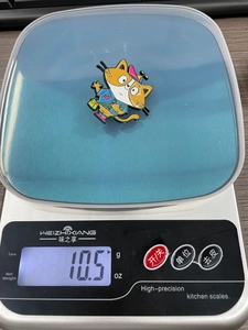 Detailed product weight measurement of cute orange cat enamel pin showing 10.5g on a scale