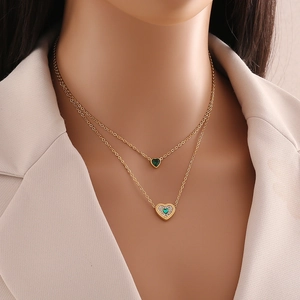 Double-layered golden necklace featuring green emerald heart pendants on a beige blazer background