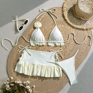 Flat lay of white three-piece bikini with 3D floral accent and ruffled skirt on straw mat