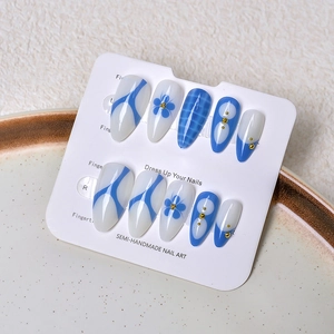 Detail of blue and white almond press-on nails with floral patterns and gold beads