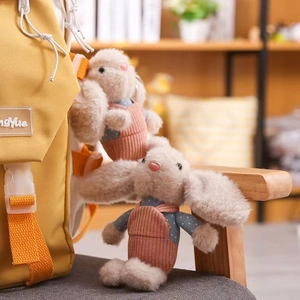 Detail of cute beige plush rabbit keychain with overalls hanging on a yellow backpack