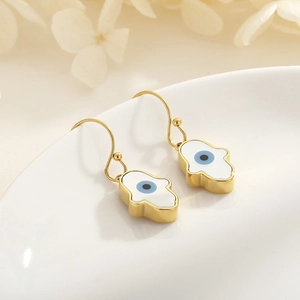 Macro shot of gold Hamsa hand earrings with blue evil eye detail on white ceramic