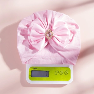 Detail of baby turban hat showing large satin bow with pink gemstone on digital scale