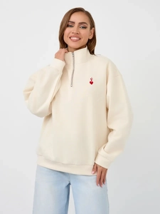 Detail of a beige half-zip sweatshirt featuring a red heart and 'V' logo embroidery