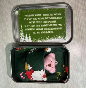 Pink teddy bear in a festive metal gift box with a Christmas card and green bedding