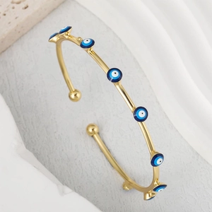 Golden open bangle bracelet featuring blue evil eye charms on a stone and fabric surface