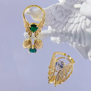Elegant gold wing rings with green and clear heart stones on a reflective surface