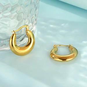 Detail of polished gold oval hoop earrings against a light blue background with glassware