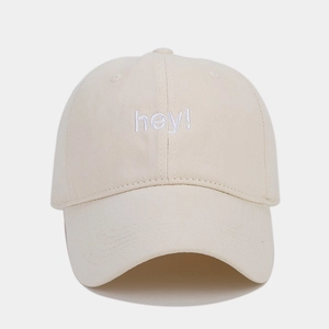 Detailed view of white 'hey!' embroidery on a beige cotton baseball cap with twill texture