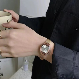 Detail view of a square gold watch with mesh strap held near a cup in a lifestyle setting