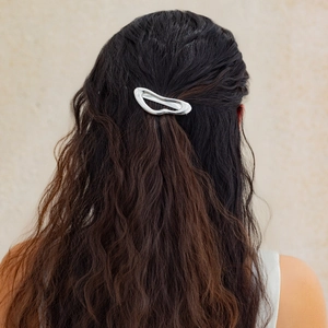Detail of silver liquid metal hair clip worn on dark wavy hair against a beige wall