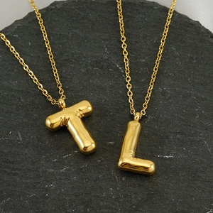3D gold bubble letter T and L pendant necklaces on grey stone surface