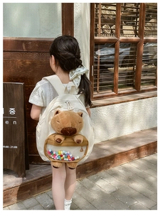 Detail of a beige kids' backpack featuring a plush capybara and clear pocket with pom-poms
