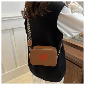 Brown leather crossbody bag detail featuring red heart motif and wide geometric strap
